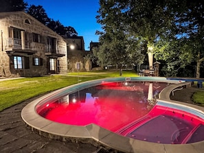 Ein Pool mit geschwungener Kante, umgeben von einem Steinhaus mit Balkon und einem Baum im Hintergrund.