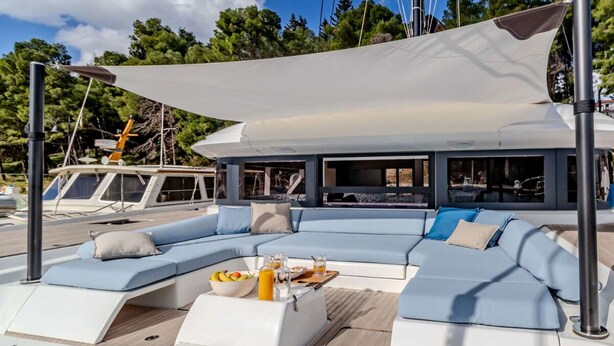 Uno yacht con una zona salotto coperta, un tavolo da pranzo con bevande e vista sul porto turistico.