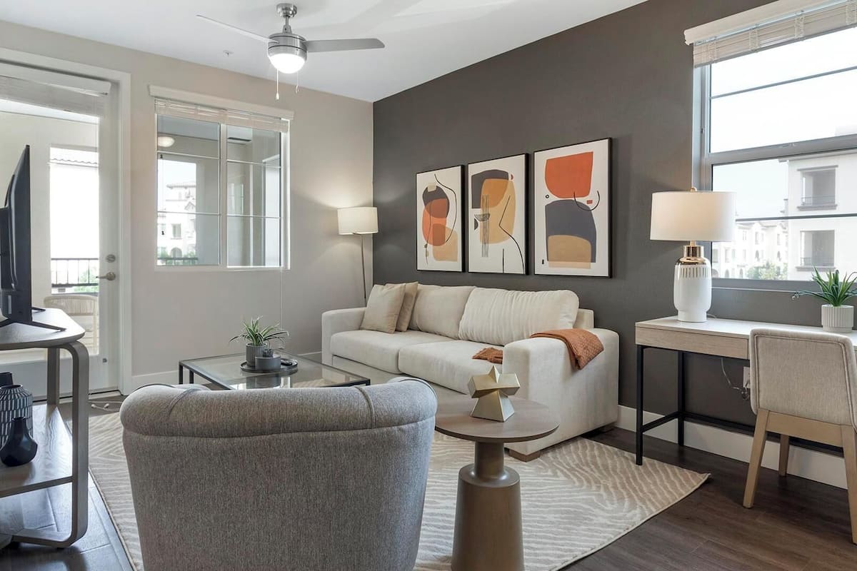 A modern living room with a sofa, coffee table, and a desk with a lamp. There are two wall-mounted artworks and a ceiling fan.