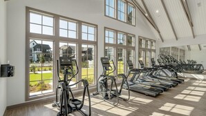 Une salle de sport équipée de plusieurs tapis de course et de plusieurs appareils elliptiques, de grandes fenêtres donnant sur un quartier résidentiel et d’un plafond haut avec des poutres en bois.