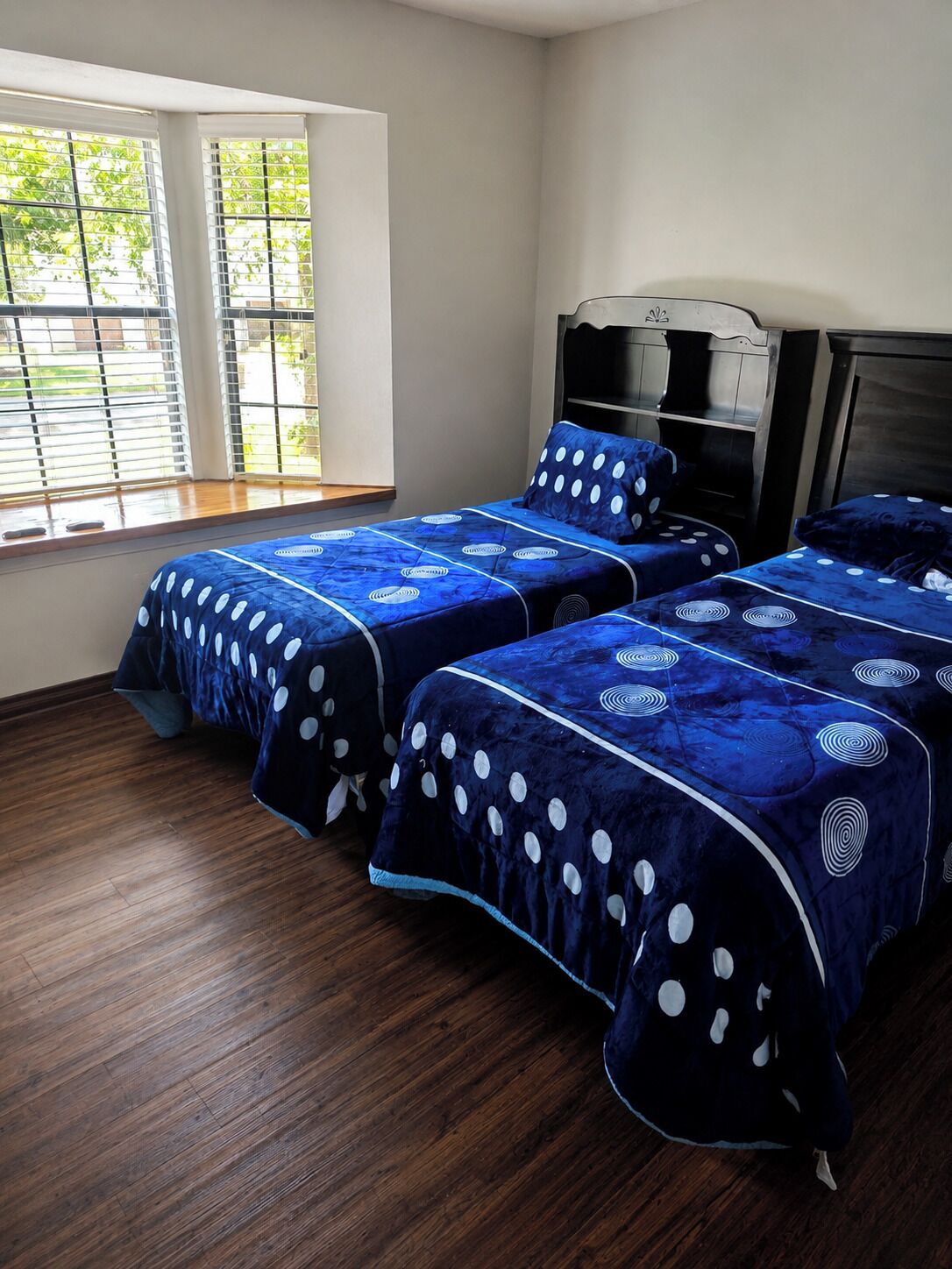 Une chambre avec deux lits, une fenêtre avec des stores et un plancher en bois.