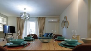 A dining area with a wooden table set for four, a television on a stand, and a ceiling light fixture. - Mediterranean 2 bedroom Luxury (Glifada)