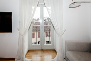 A room with a balcony, white curtains, a grey sofa, and a TV on the wall. - Nuccia Mansarda in heart of Como  (COMO)