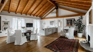 Ein geräumiges Wohnzimmer mit Holzbalken, einem Essbereich mit weißen Stühlen, einer Couch, einem Kamin und einer großen Pflanze.