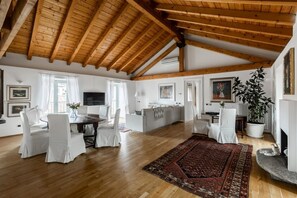 Ein geräumiges Wohnzimmer mit Holzbalken, einem Essbereich mit weißen Stühlen, einer Couch, einem Kamin und einer großen Pflanze.