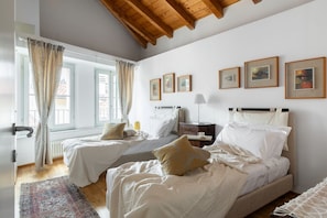 A bedroom with two beds, a window with curtains, a radiator, and framed pictures on the wall. - Nuccia Mansarda in heart of Como  (COMO)