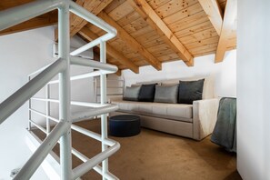 Ein Loft-Zimmer mit Holzbalkendecke, einer weißen Treppe, einem beigen Sofa mit Kissen und einem runden Hocker.