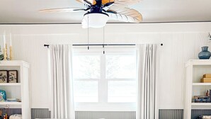 Ett vardagsrum med en soffa, ett soffbord i glas, en takfläkt och ett fönster med gardiner.