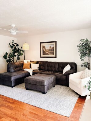 Un salon comprenant un canapé d’angle, un fauteuil blanc, une grande plante en pot, un ventilateur de plafond et un tableau encadré accroché au mur.