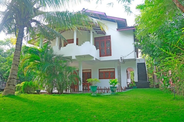 Une maison Ă deux Ă©tages dotĂ©e dâun balcon, entourĂ©e dâune vĂ©gĂ©tation luxuriante et de palmiers.