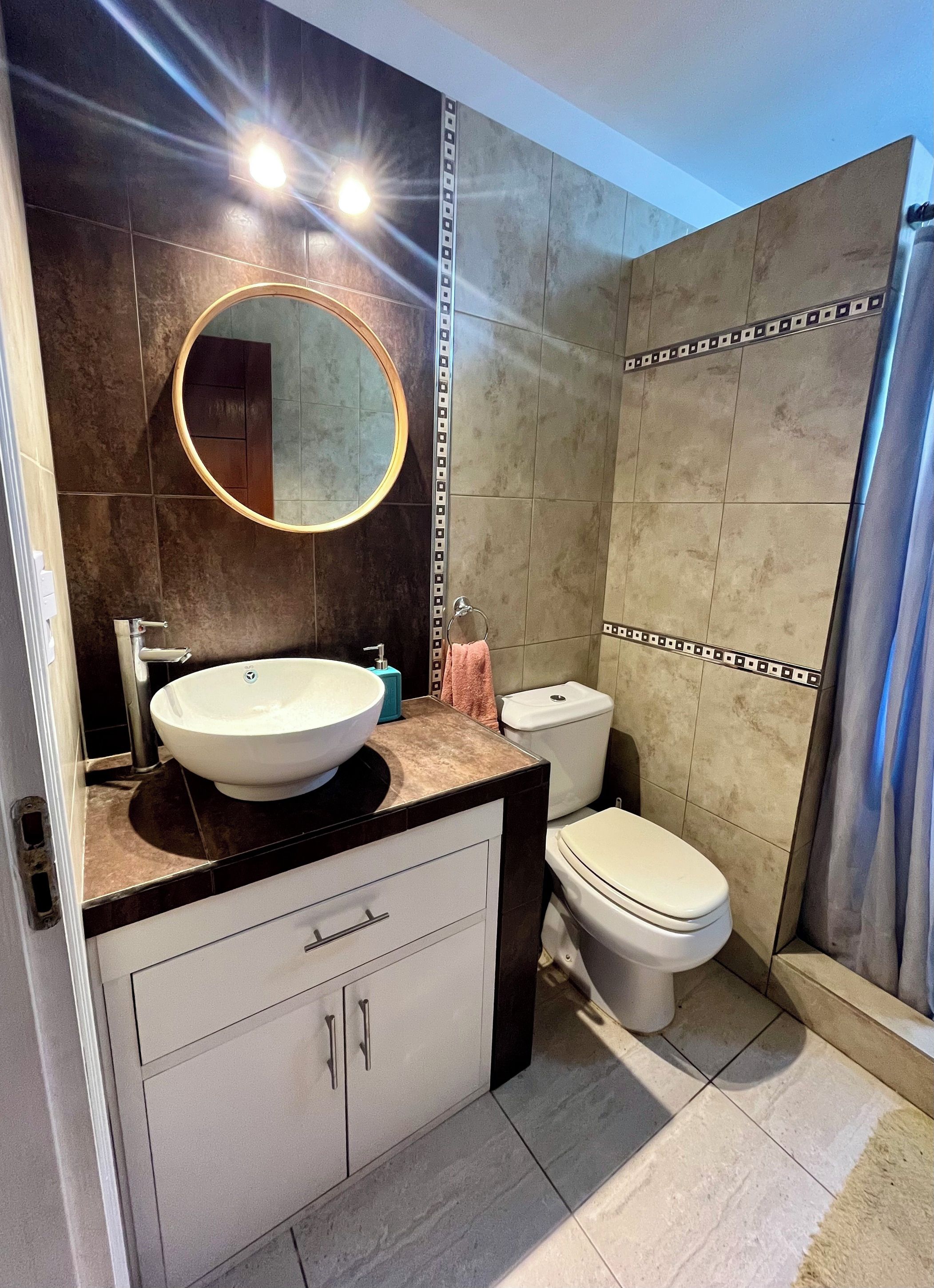 Une salle de bain avec un miroir rond, un lavabo blanc, des toilettes et un rideau de douche.