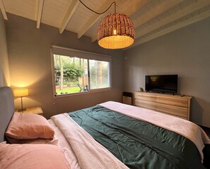 A bedroom with a bed, a dresser, a television, a lamp, and a window with a view of the outdoors.