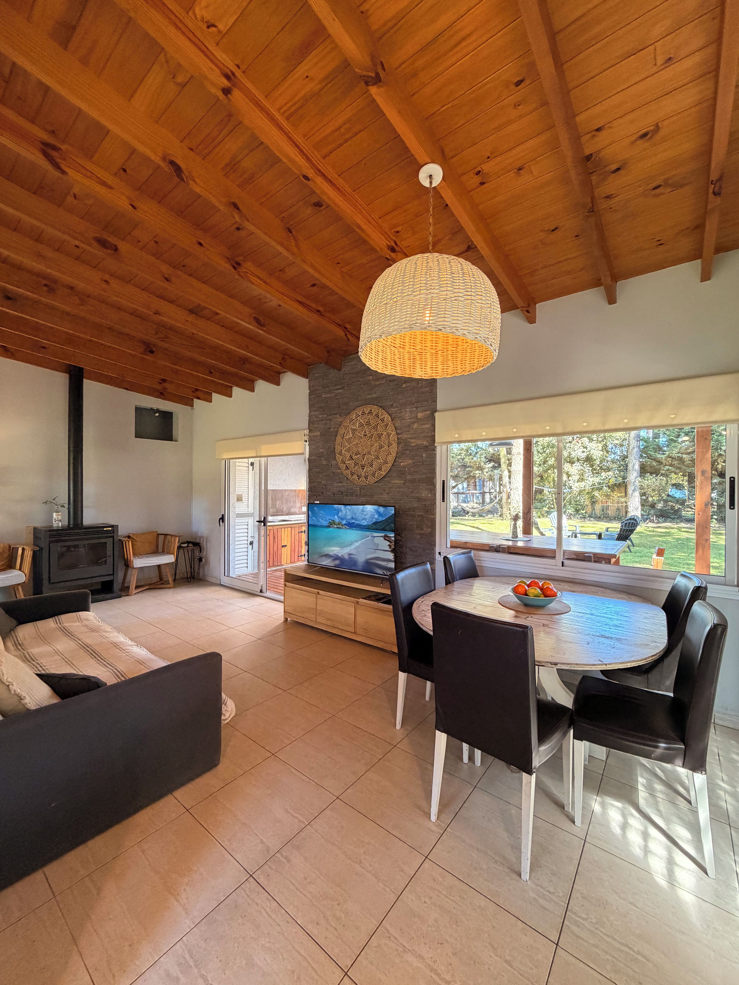 A spacious living area with a wooden ceiling, a fireplace, a dining table with chairs, and a large window offering a view of the outdoors.