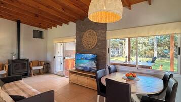 A spacious living area with a wooden ceiling, a fireplace, a dining table with chairs, and a large window offering a view of the outdoors.