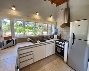 A modern kitchen with a stove, sink, and refrigerator, large windows with a view of greenery, and a water dispenser.