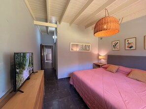 Une chambre avec un grand lit, un téléviseur sur le côté gauche, un couloir menant à une autre pièce, et un plafond avec des poutres apparentes.