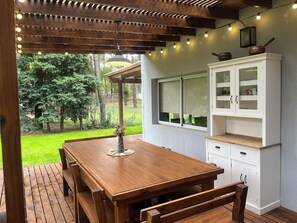 Une table à manger en bois avec des bancs, un buffet blanc avec des portes en verre et une pergola illuminée de guirlandes lumineuses.