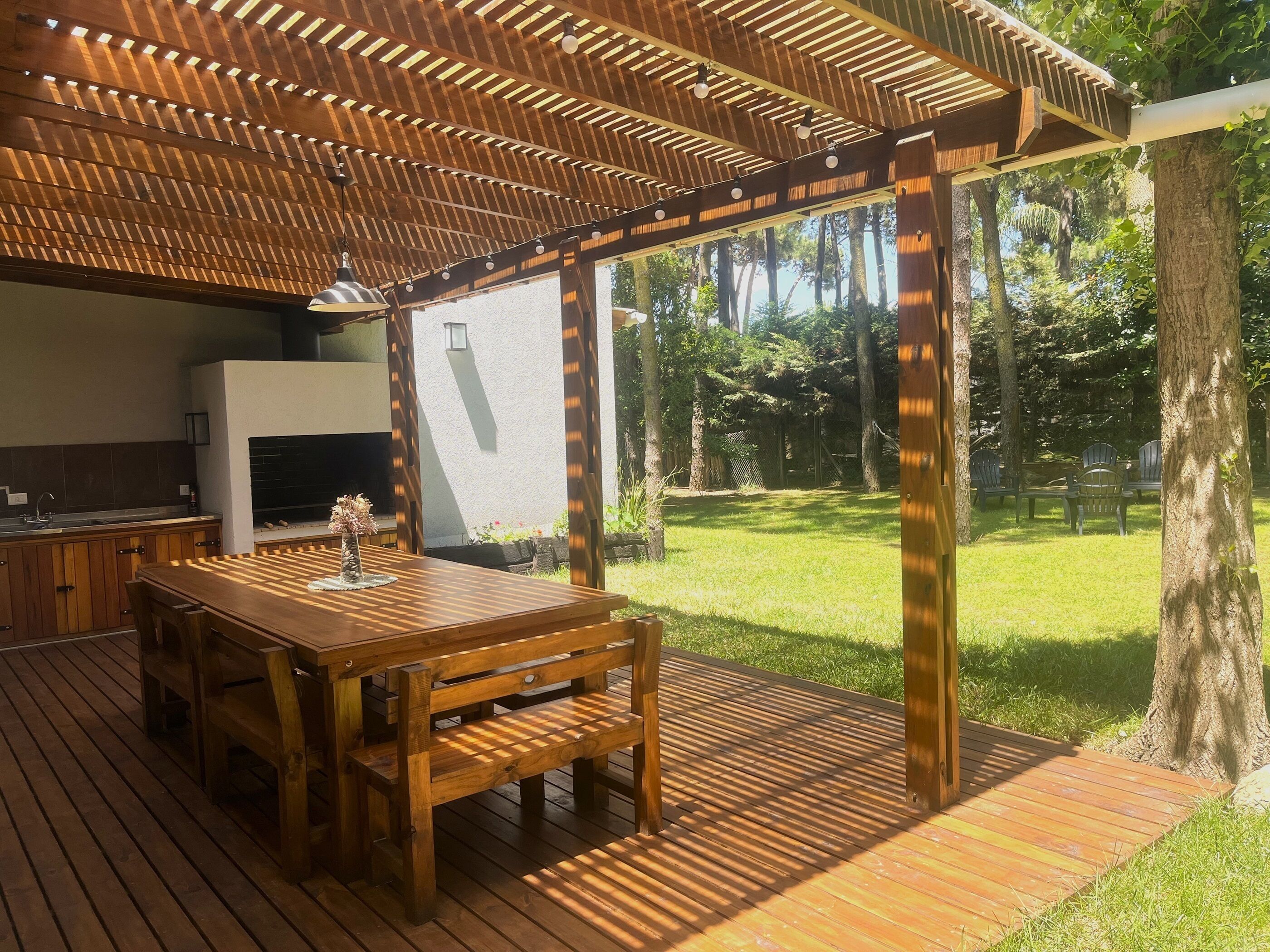 Une terrasse en bois avec une table et des chaises, un barbecue et une pergola illuminée de guirlandes lumineuses.
