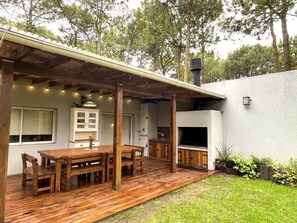 A wooden deck with a dining table and chairs, a built-in grill, and a white exterior wall.