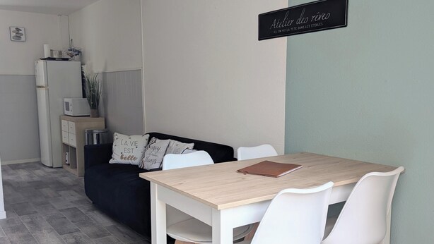 Un comedor con una mesa de madera y sillas blancas, un sofá negro con cojines decorativos, una pequeña cocina con microondas y un cartel en la pared.