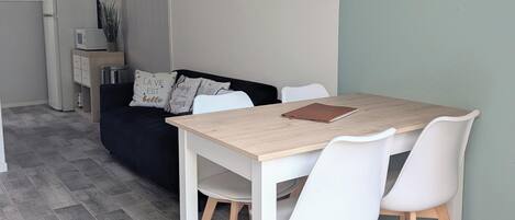 A dining area with a wooden table and white chairs, a black couch with decorative pillows, a small kitchen area with a microwave, and a sign on the wall.
