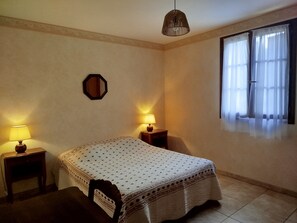 A bedroom with a bed, two bedside lamps, a hanging light fixture, a window with curtains, and a mirror on the wall. - Gîte Candéjac (Daglan)