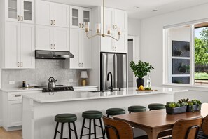 A modern kitchen with white cabinetry, a stainless steel refrigerator, a gas stove, and a central island with bar stools. - New! Lake LBJ Waterfront l 5BR Luxury Home (Kingsland)