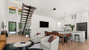 A modern living room with a staircase, a dining area, and a kitchen with white cabinetry and a black refrigerator.