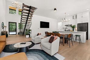 A modern living room with a staircase, a dining area, and a kitchen with white cabinetry and a black refrigerator. - New! Lake LBJ Waterfront l 5BR Luxury Home (Kingsland)