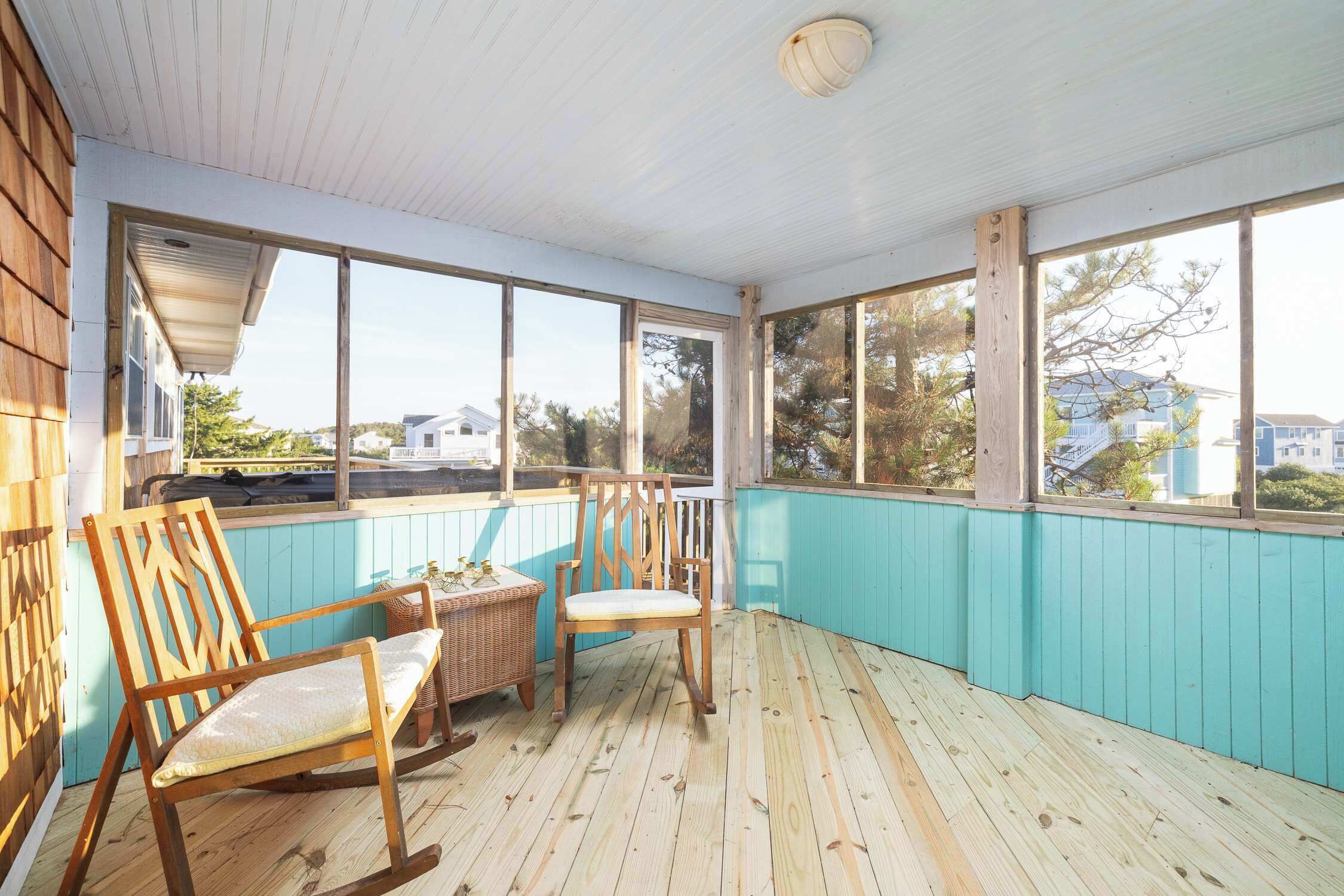 Une véranda fermée avec un plancher en bois, deux chaises en bois, une table en osier et une vue sur les maisons et les arbres.