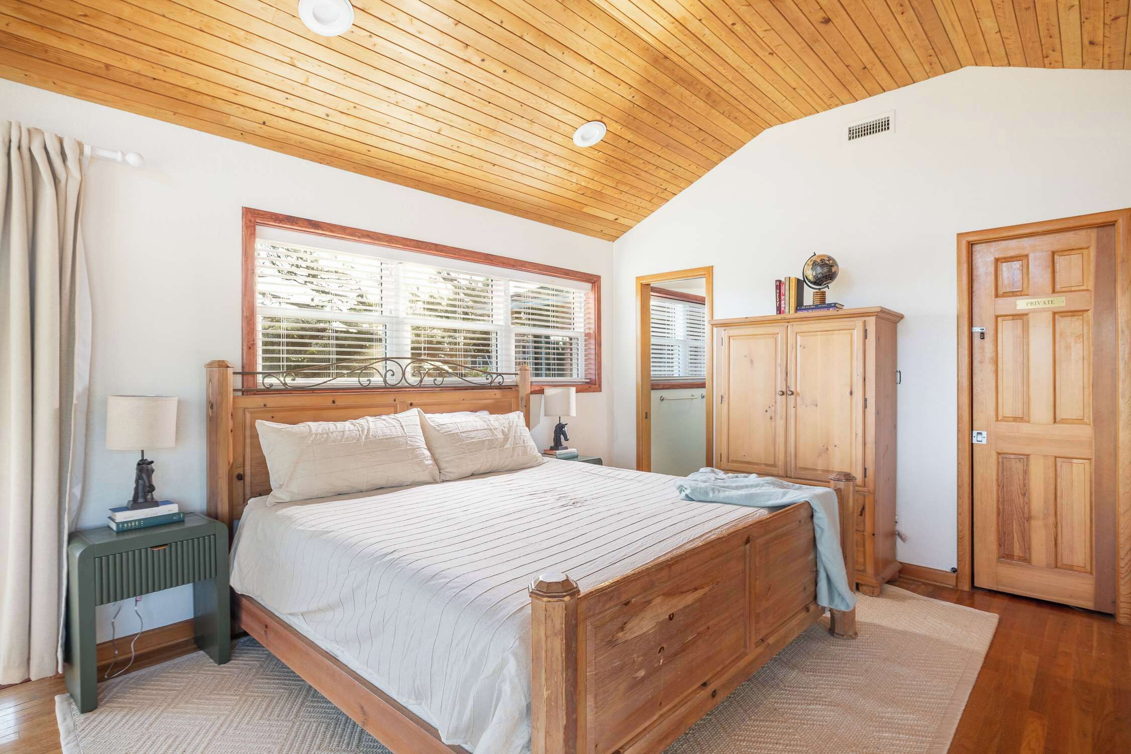 A bedroom with a wooden ceiling, a large bed, a nightstand, a mirror, a wardrobe, and a window with blinds.