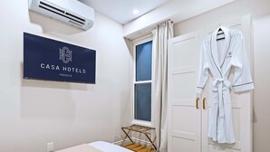 A hotel room with a bed, a robe hanging on a door, a window with curtains, an air conditioning unit, and a sign with the hotel's name. - The Gerrard | Group Basement CN Tower Transit Base (Toronto)