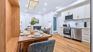 A modern kitchen with white cabinets, stainless steel appliances, and a dining area with a wooden table and chairs. - The Gerrard | Group Basement CN Tower Transit Base (Toronto)
