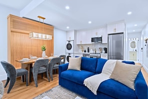 A modern kitchen with white cabinets, a stainless steel refrigerator, and a dining area with a wooden table and grey chairs.