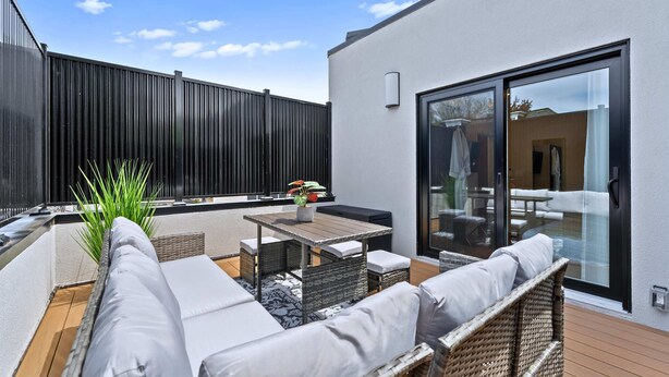 Un patio moderno al aire libre con mobiliario de mimbre, una mesa de madera y una puerta corredera de vidrio que da a un espacio interior.