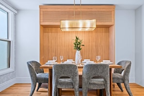 A dining area with a wooden table, four grey upholstered chairs, a white vase with greenery, and a pendant light.