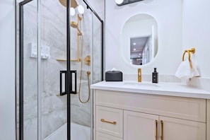 A modern bathroom with a glass-enclosed shower, a white marble wall, a round mirror, and a beige vanity with gold handles.