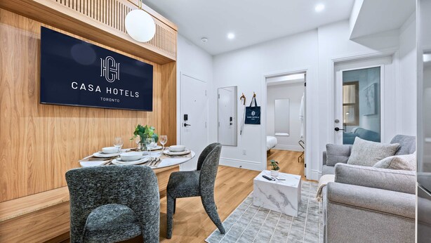 Moderni hotellihuone, jossa on ruokailutila, sohva ja televisio, jossa näkyy hotellin nimi.