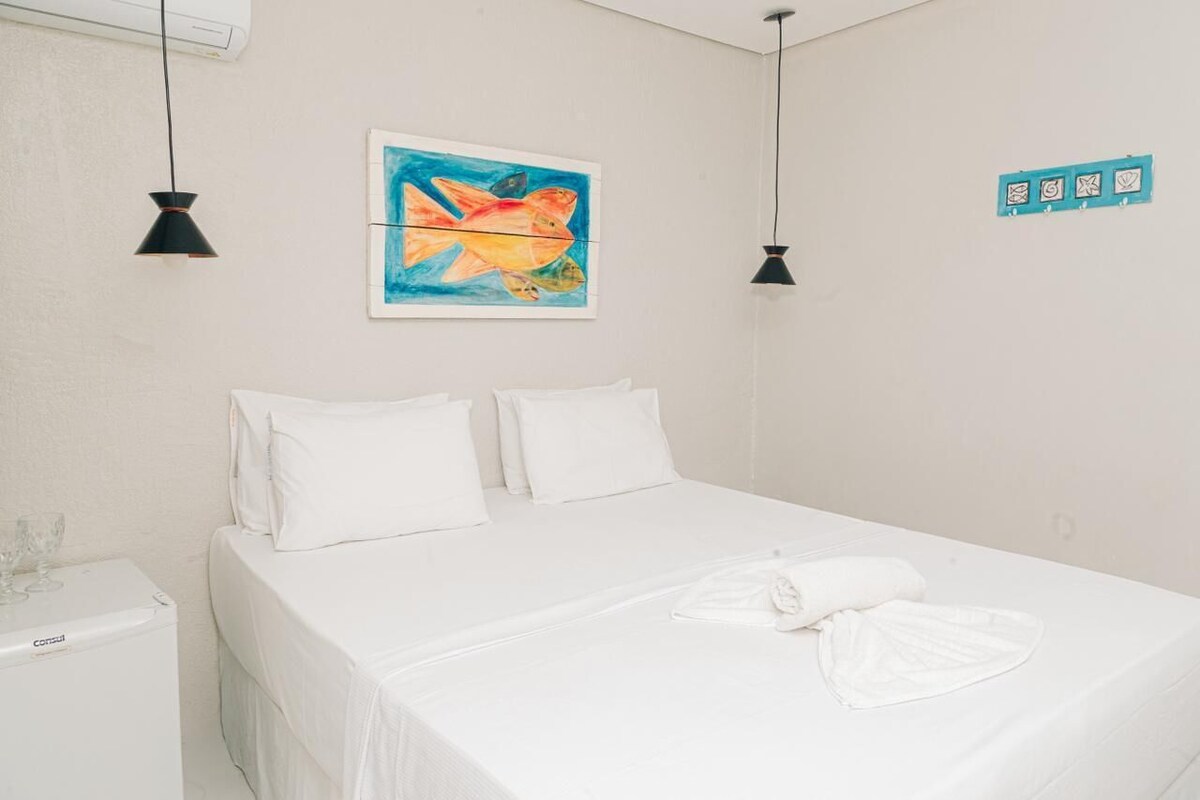 A hotel room with a bed, white bedding, a painting, two pendant lights, and a small table with a glass.