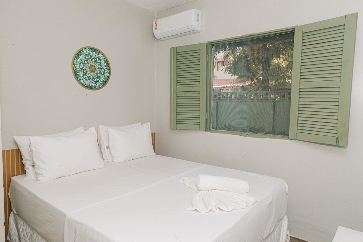 A hotel room with a bed, pillows, a window with shutters, and a decorative plate on the wall.