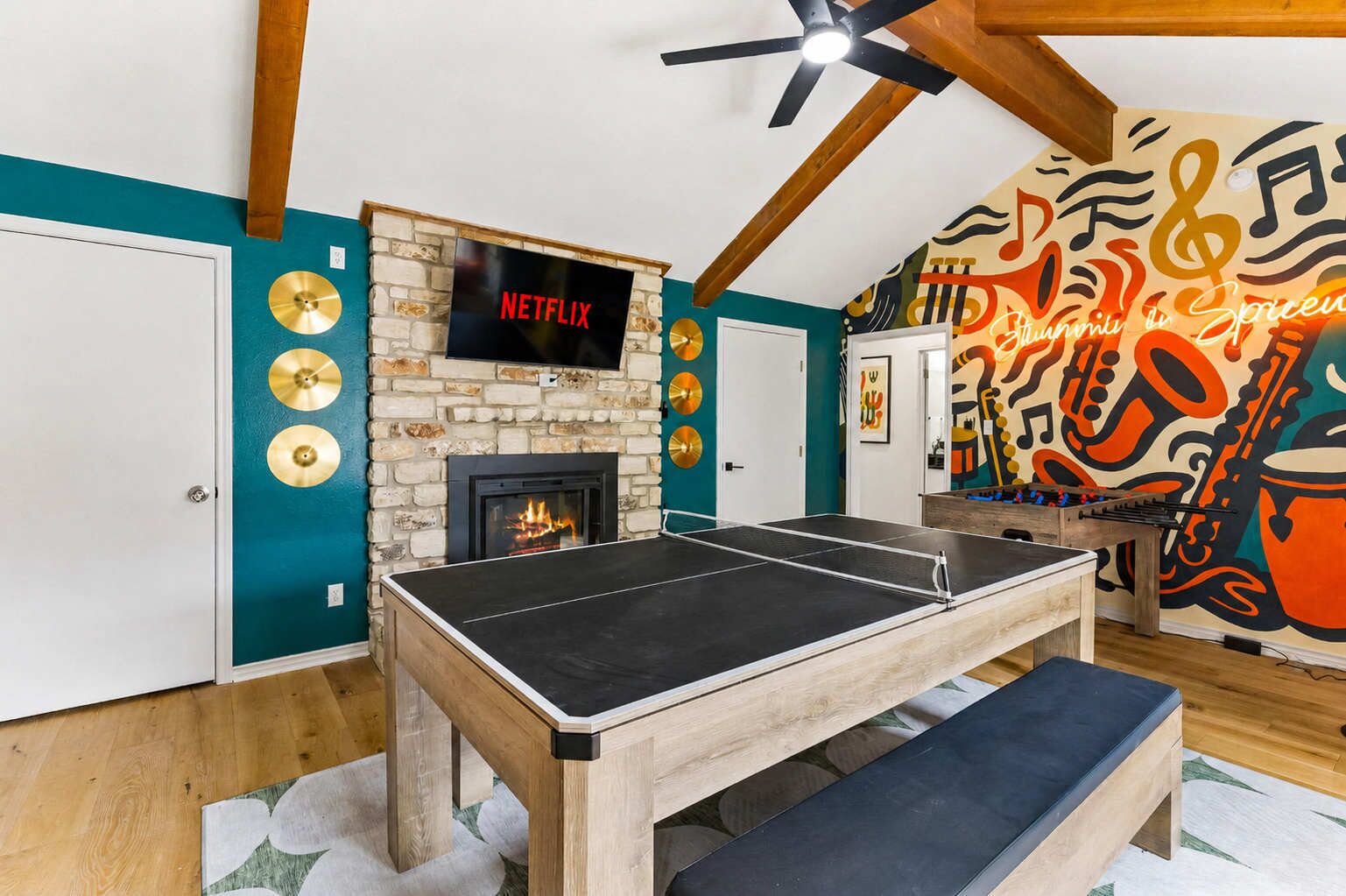 A room with a ping pong table, a fireplace, a TV mounted on a stone wall, and a colorful mural.