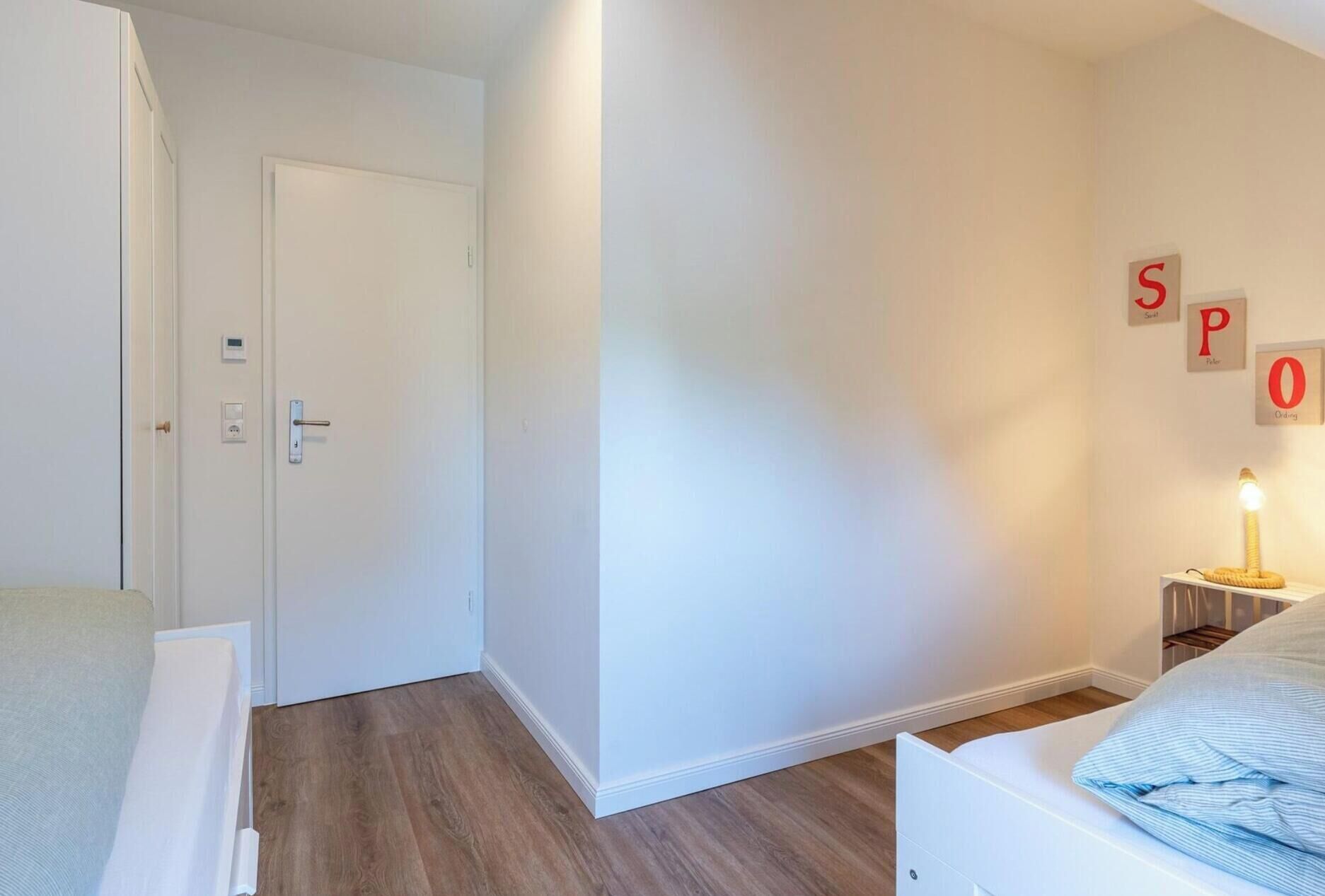 Une chambre à coucher avec un lit, une table de chevet avec une lampe, une porte dotée d’un thermostat et un mur sur lequel sont inscrites les lettres formant le mot « SPOO ».