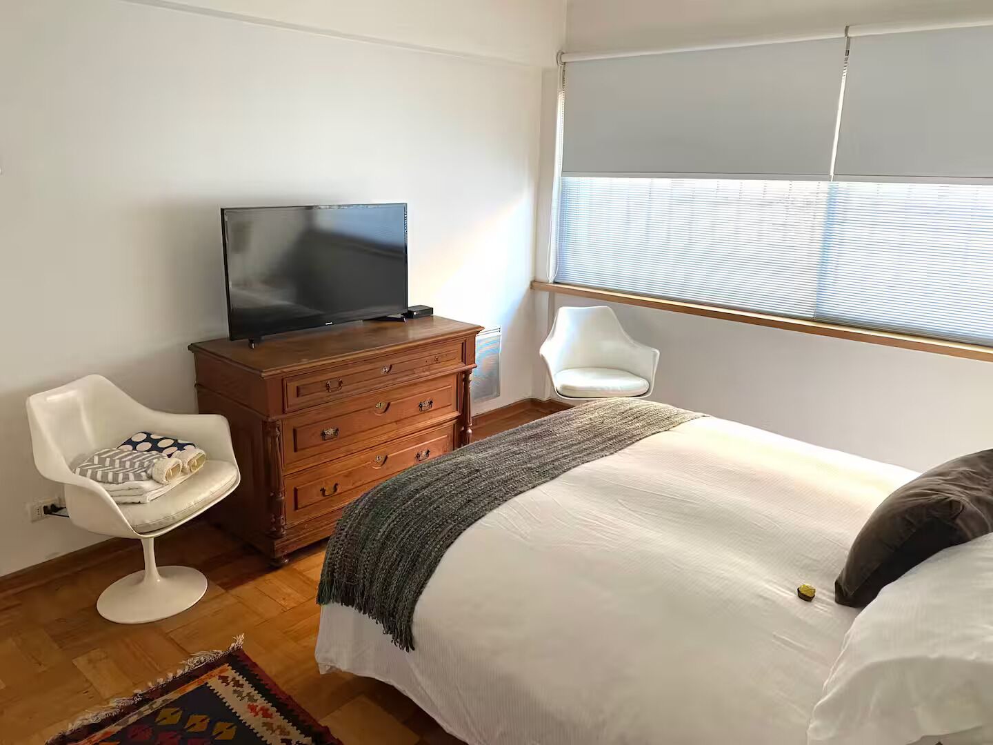 Ein Schlafzimmer mit einem Bett, einem Fernseher auf einer Holzkommode, einem weißen Stuhl und einem Fenster mit Jalousien.
