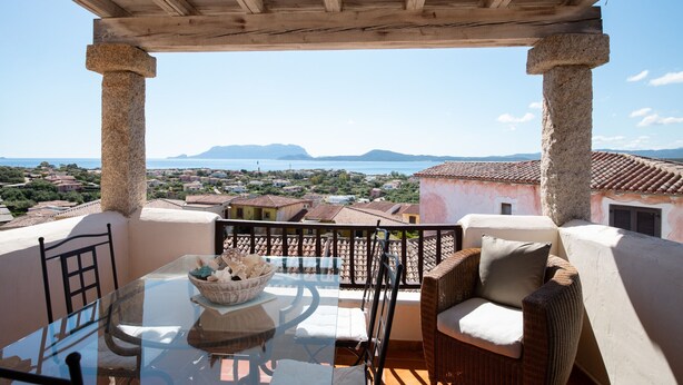 A balcony with a glass table and wicker furniture, overlooking a coastal town and mountains. - Casa dell'Erica by Wonderful Italy (Olbia)