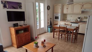A compact living space with a dining table, chairs, a TV, a wooden cabinet, and a kitchen area with cabinets and appliances. - The Bicentennial Linden Cottage (Pernes-les-Fontaines)