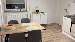 Cocina compacta con una mesa, sillas, ventana con persianas, radiador y un mueble pequeño.