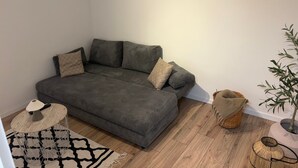 Una habitación de esquina con un sofá gris, cojines estampados, una mesa redonda de madera con una lámpara, una alfombra tejida, una cesta de mimbre y una planta en maceta.