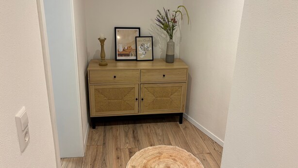 Una habitación de esquina con un armario de madera, un cuadro enmarco, un florero con flores y una alfombra tejida.