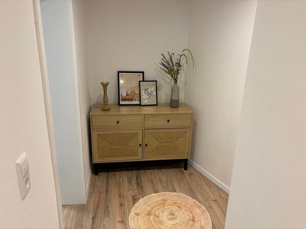 A corner room with a wooden cabinet, a framed picture, a vase with flowers, and a woven rug.