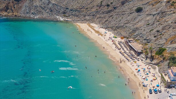 Une plage côtière aux eaux turquoise et limpides, bordée d’un rivage sablonneux et d’une falaise rocheuse.